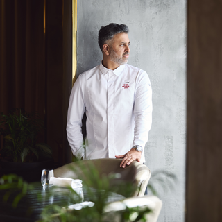 A Michelin-starred chef wearing a white long-sleeve chef coat with a Michelin logo on the left chest stands in a restaurant setting, leaning on a chair near a window.