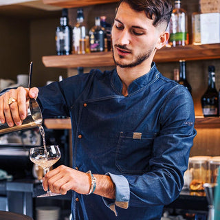 A man in a dark blue denim-style chef jacket pouring a drink in a bar setting.