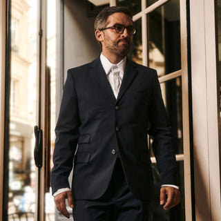 A maître d' wearing a dark navy blue two-piece suit, white dress shirt, and glasses standing in front of a glass door.