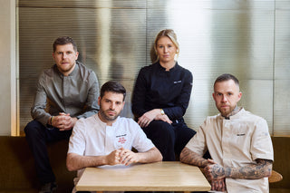 Four executive chefs in uniforms posing together in front of a modern slatted metal wall. 