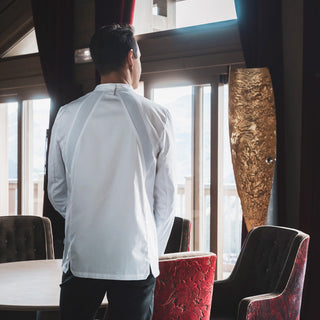 Chef wearing a classic white long-sleeve men's chef coat with a mandarin collar and a central front fastening. It features elasticated panels on the upper back and a loop on the back collar for securing an apron.