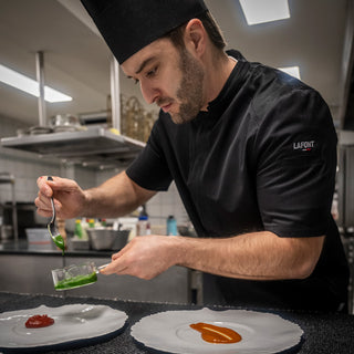 Kitchen chef, plating a dish in a black, short-sleeve men's chef coat with a mandarin collar and a front pocket.